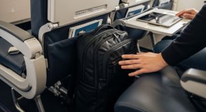 A traveler sliding a slim black backpack under an airplane seat in an economy cabin
